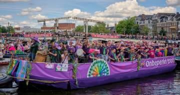 Paarse boot met mensen die zwaaien, aan de Amsterdamse kade zijn veel zwaaiende mensen te zien