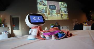 Robot Ivy staat met een gebreide sjaal op tafel, daarachter staat de eerste slide van de presentatie die gegeven werd bij de feestelijke afronding van het programma sociale robotica